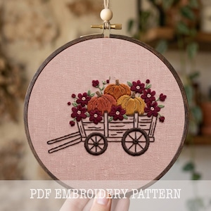 Op de afbeelding: Een ronde houten borduurring met een oudroze stoffen paneel met een handgestikt ontwerp van een houten kar gevuld met oranje en gele pompoenen, omringd door donkerrode bloemen. De tekst PDF EMBROIDERY PATTERN is zichtbaar op de afbeelding.