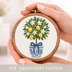 May include: A detailed embroidery of a lemon tree in a blue and white pot, set within a wooden hoop. The tree features yellow lemons, white flowers, and green leaves. The text "PDF EMBROIDERY PATTERN" is at the bottom.