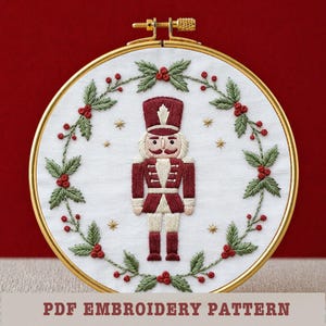 Puede incluir: Arte de aro bordado con un soldado cascanueces en uniforme burdeos, rostro blanco y pantalón beige. El diseño está rodeado por una corona de hojas verdes y bayas rojas, con estrellas doradas sobre un fondo blanco. El texto "PDF EMBROIDERY PATTERN" está en la parte inferior.