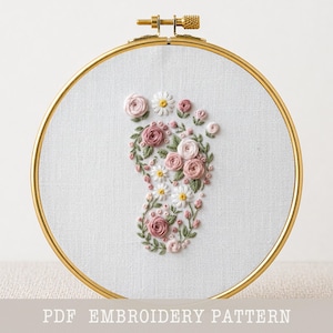 May include: A circular gold embroidery hoop containing white fabric with a hand-stitched floral design in the shape of a baby footprint. The embroidery features pink roses, small white daisies, and green leaves. The text PDF EMBROIDERY PATTERN is visible.