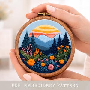Könnte beinhalten: Ein runder Stickrahmen mit einer Landschaftsszene mit Sonnenuntergang, Bergen und Bäumen. Die Stickerei verwendet blaue, orange und grüne Farben. Der Text "PDF EMBROIDERY PATTERN" ist unten zu sehen.