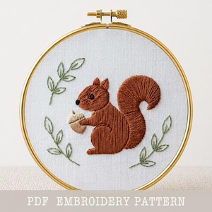 May include: Embroidered squirrel holding an acorn, framed in a gold hoop. The squirrel is brown with a fluffy tail, surrounded by green leaves on a white background. Text at the bottom reads "PDF EMBROIDERY PATTERN".