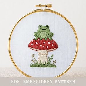 May include: An embroidered design in a gold hoop. A green frog is perched on a red mushroom with white spots. The embroidery is set against a white fabric, with the text "PDF EMBROIDERY PATTERN" at the bottom.