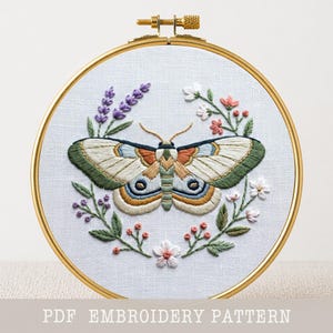 May include: Embroidered moth design in a gold hoop. The moth has cream-colored wings with green, blue, and orange details. Lavender, small white, and pink flowers surround the moth. The text "PDF EMBROIDERY PATTERN" is at the bottom.