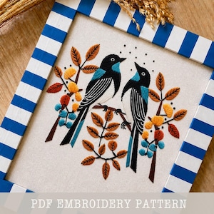 May include: Embroidered artwork featuring two birds with black and white bodies and turquoise wings, perched on a branch with autumn-colored leaves and flowers. The artwork is framed in a blue and white striped wooden frame. The text "PDF EMBROIDERY PATTERN" is at the bottom.