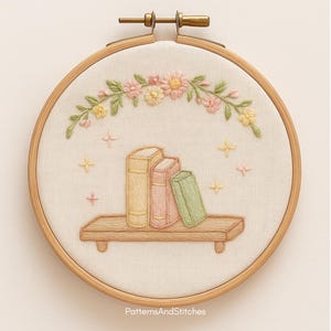 Puede incluir: Arte bordado en bastidor que muestra una estanter&iacute;a con tres libros en colores pastel. Una estanter&iacute;a de madera sostiene los libros, con un arco floral encima. El fondo es de tela beige claro, y el bastidor es de madera. El texto "PatternsAndStitches" est&aacute; en la parte inferior.