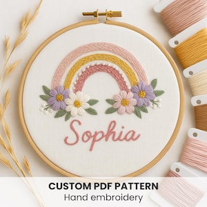 Puede incluir: Un bordado a mano en bastidor con un diseño de arcoíris en tonos rosa y amarillo, detalles florales y el nombre "Sophia" bordado en rosa. El texto "CUSTOM PDF PATTERN Hand embroidery" está en la parte inferior.