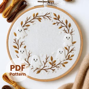 Pode incluir: Arte de bordado em bastidor com tecido branco com um desenho costurado de figuras de fantasmas sorridentes e folhas de outono. O texto "PDF Pattern" é visível. O bastidor de madeira é rodeado por fios de bordar castanhos e amarelos.