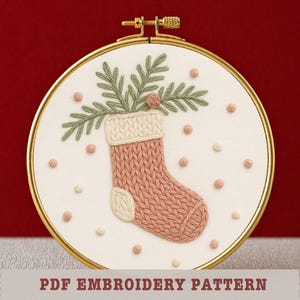 May include: A round embroidery hoop with a gold-colored frame. The design features a pink stocking with a cream-colored cuff, green sprigs, and small pink and cream-colored dots. The text "PDF EMBROIDERY PATTERN" is at the bottom.