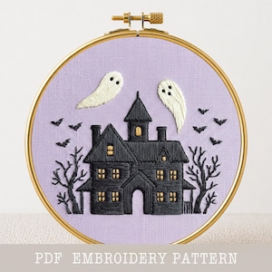 May include: A circular embroidery hoop containing a lavender fabric background with a hand-stitched design of a black haunted house, two white ghosts, black bats, and bare trees. The text PDF EMBROIDERY PATTERN is printed at the bottom.