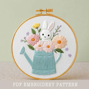 May include: Embroidered artwork in a gold hoop features a white bunny peeking out from a blue watering can filled with colorful flowers. The flowers include pink, yellow, and peach blooms. The text "PDF EMBROIDERY PATTERN" is at the bottom.