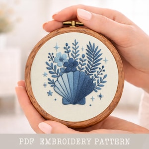 Könnte beinhalten: Ein runder Holz-Stickrahmen mit einem blauen Muscheldesign und floralen Akzenten. Das Design ist auf cremefarbenem Stoff gestickt. Der Text "PDF EMBROIDERY PATTERN" befindet sich am unteren Rand des Bildes.