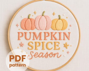 May include: An embroidery hoop containing white fabric with hand-stitched embroidery of three pumpkins in peach, light pink, and yellow. The text reads PUMPKIN SPICE Season in orange and yellow thread, accented by small embroidered stars and flowers.