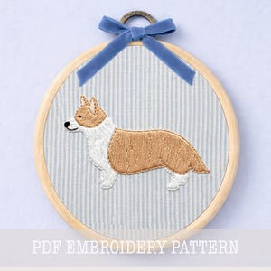 Può includere: Corgi ricamato su sfondo a righe, in un cerchio di legno. Il corgi è marrone e bianco, con un nastro blu in alto. Il testo "PDF EMBROIDERY PATTERN" è in basso.