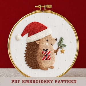以下が含まれることがあります： サンタ帽をかぶったクリスマスのハリネズミの刺繍。松の小枝と包まれたプレゼントを持っています。デザインは金色の刺繍枠にセットされています。「PDF EMBROIDERY PATTERN」の文字が下部にあります。