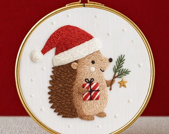 Könnte beinhalten: Gestickter Weihnachts-Igel mit Weihnachtsmütze, der einen Tannenzweig und ein Geschenk hält. Das Design befindet sich in einem goldfarbenen Stickrahmen. Der Text "PDF EMBROIDERY PATTERN" steht unten.