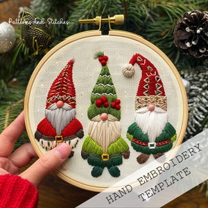 May include: Three embroidered gnomes in a wooden embroidery hoop. The gnomes are wearing red, green and white hats and have white beards. The text "HAND EMBROIDERY TEMPLATE" is printed below the hoop.