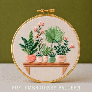Puede incluir: Arte bordado en un aro dorado que representa plantas en macetas sobre una estantería de madera. Las plantas son de varios tonos de verde y rosa, sobre un fondo beige. El texto "PDF EMBROIDERY PATTERN" es visible en la parte inferior.