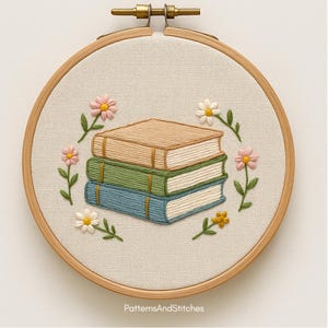 Puede incluir: Arte bordado en un aro de madera. Muestra una pila de tres libros en beige, verde y azul, rodeada de flores rosas y blancas y hojas verdes. El dise&ntilde;o est&aacute; sobre tela crema. El texto "PatternsAndStitches" est&aacute; en la parte inferior.