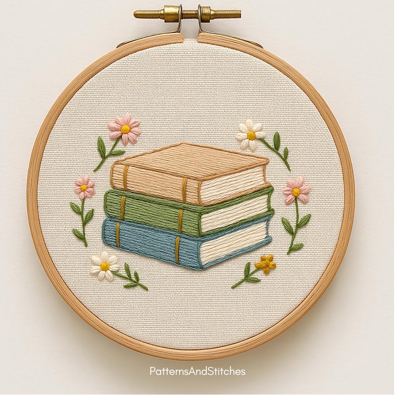 Puede incluir: Arte bordado en un aro de madera. Muestra una pila de tres libros en beige, verde y azul, rodeada de flores rosas y blancas y hojas verdes. El dise&ntilde;o est&aacute; sobre tela crema. El texto "PatternsAndStitches" est&aacute; en la parte inferior.