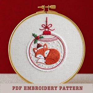 May include: Embroidered Christmas ornament featuring a sleeping fox wearing a Santa hat. The fox is orange and white, inside a white and red ornament design. The embroidery is set in a gold hoop. Text at the bottom reads "PDF EMBROIDERY PATTERN".