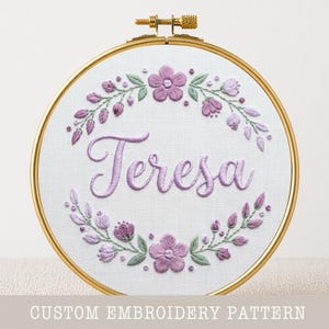 May include: Embroidered hoop art with the name "Teresa" in lavender script, encircled by a floral wreath of lavender and green embroidered flowers and leaves. The hoop is gold. Text at the bottom reads "CUSTOM EMBROIDERY PATTERN".