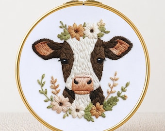 May include: Embroidered cow face with a floral crown and surrounding greenery, set in a gold hoop. The cow has brown and white markings, with detailed stitching. Text at the bottom reads "PDF EMBROIDERY PATTERN".
