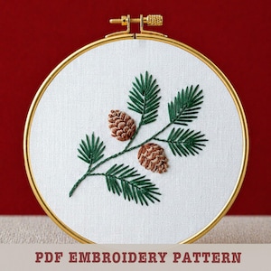 May include: Embroidered pinecone and evergreen branch design on white fabric, set in a gold hoop. The embroidery features brown pinecones and green needles. The text "PDF EMBROIDERY PATTERN" is at the bottom.