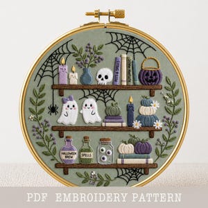 Puede incluir: Un bastidor de bordado circular con tela verde salvia y tres estantes de madera bordados en hilo marrón. Los estantes muestran elementos bordados que incluyen fantasmas, calabazas, calaveras, velas, libros etiquetados Pots & Spells y Autumn Magic, y frascos etiquetados Halloween Brew y Spells, rodeados de telarañas y enredaderas florales.