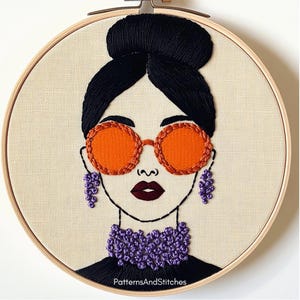 May include: Embroidered portrait of a woman in a wooden hoop. The woman has black hair in a bun, orange sunglasses, and purple earrings and necklace. The text "PatternsAndStitches" is at the bottom.