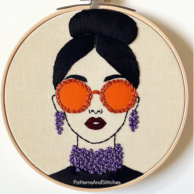 May include: Embroidered portrait of a woman in a wooden hoop. The woman has black hair in a bun, orange sunglasses, and purple earrings and necklace. The text "PatternsAndStitches" is at the bottom.