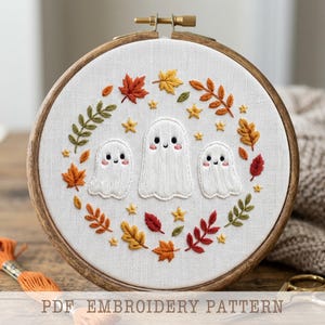 May include: An embroidery hoop containing white fabric with a hand-stitched design of three white ghosts surrounded by a wreath of colorful autumn leaves and small yellow stars. The text PDF EMBROIDERY PATTERN is printed at the bottom.