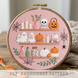May include: A circular wooden embroidery hoop containing a pink fabric background with three shelves stitched in brown thread. The shelves display embroidered items including white ghosts, orange and white pumpkins, candles, a skull, and jars.