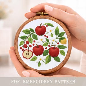 May include: A round embroidery hoop featuring a detailed floral design with red apples, white flowers, green leaves, and a yellow lemon. The embroidery is set against a white background and framed by a wooden hoop. The text "PDF EMBROIDERY PATTERN" is at the bottom.