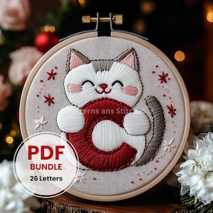May include: Embroidered hoop art featuring a white cat holding a red letter C. The cat has brown ears and a brown tail. The background is a light beige fabric with white and red stars.