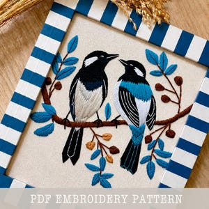 May include: Embroidered artwork featuring two birds perched on a branch with blue leaves and brown berries. The birds have black and white plumage with blue accents. The artwork is framed in a blue and white striped frame. The text "PDF EMBROIDERY PATTERN" is at the bottom.