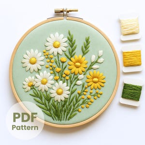 Puede incluir: Un bastidor de bordado con un diseño floral de margaritas blancas y amarillas, hojas verdes y detalles amarillos sobre un fondo verde claro. La imagen incluye tres carretes de hilo y el texto "PDF Pattern".