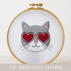 Può includere: Gatto ricamato con occhiali da sole a forma di cuore in un cerchio dorato. Il gatto è grigio con accenti rosa e occhiali da sole rossi. Il testo "PDF EMBROIDERY PATTERN" è in basso.