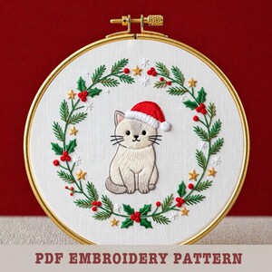 May include: Embroidered cat wearing a Santa hat, centered in a wreath of green leaves, red berries, and gold stars. The design is set in a gold hoop. Text at the bottom reads "PDF EMBROIDERY PATTERN".