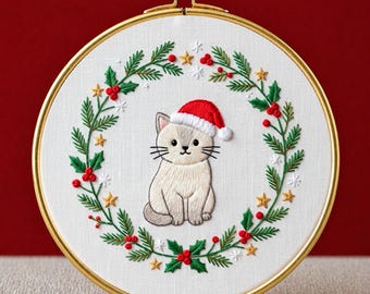 May include: Embroidered cat wearing a Santa hat, centered in a wreath of green leaves, red berries, and gold stars. The design is set in a gold hoop. Text at the bottom reads "PDF EMBROIDERY PATTERN".