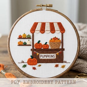 Op de afbeelding: Een ronde houten borduurring met witte stof waarop een handgeborduurd ontwerp van een pompoenkraam staat. De afbeelding toont een bruine kraam met een oranje-witte gestreepte luifel, planken met kleine pompoenen en de tekst PUMPKINS.