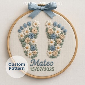 May include: Embroidered baby feet design in a wooden hoop. The design features blue, white, and tan flowers and leaves, with the name "Mateo" and the date "15/07/2025" embroidered in blue thread. A blue ribbon bow is at the top.