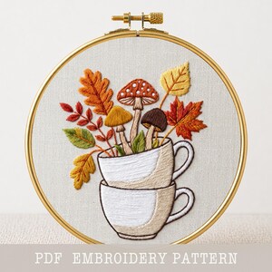 May include: An embroidery hoop containing a fabric panel with a stitched design of two stacked white teacups filled with colorful autumn leaves and three mushrooms in shades of brown, orange, and yellow. The text PDF EMBROIDERY PATTERN is visible.