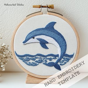 Puede incluir: Plantilla de bordado a mano con un delfín azul y blanco saltando sobre las olas. El diseño está cosido sobre tela blanca, dentro de un bastidor de bordado de madera. El texto "HAND EMBROIDERY TEMPLATE" es visible.