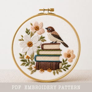 以下が含まれることがあります： 鳥が本の上に止まり、花と葉に囲まれた刺繍フープアート。白い花、桃色の花、緑の葉が含まれています。「PDF EMBROIDERY PATTERN」の文字が下にあります。