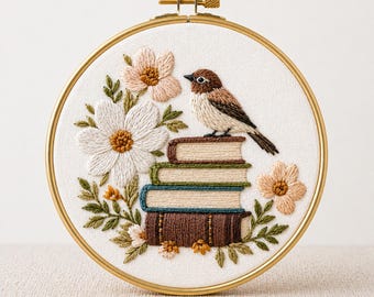 Puede incluir: Arte de aro bordado con un pájaro posado sobre una pila de libros, rodeado de flores y follaje. El diseño incluye una flor blanca, flores color melocotón y hojas verdes. El texto "PDF EMBROIDERY PATTERN" está en la parte inferior.