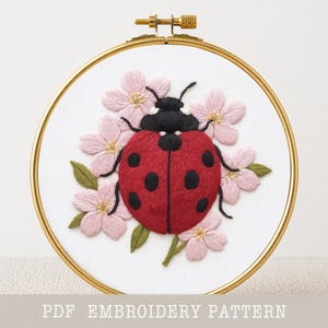 May include: Embroidered ladybug design in a gold hoop. The ladybug is red with black spots, surrounded by pink flowers and green leaves. The text "PDF EMBROIDERY PATTERN" is at the bottom.