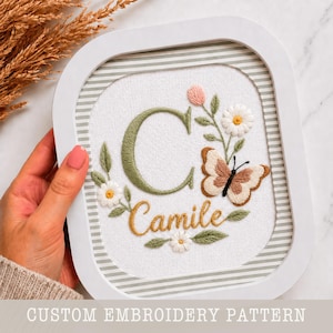 May include: Embroidered artwork in a white frame. The design features a green letter "C", the name "Camile" in gold, a butterfly, and floral accents. The frame has a striped border. The text at the bottom reads "CUSTOM EMBROIDERY PATTERN".
