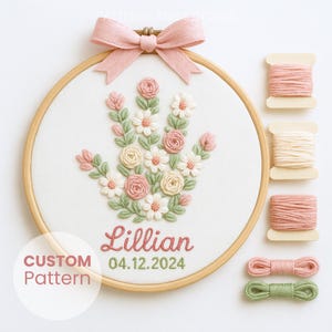 以下が含まれることがあります： ピンクのリボンが付いた木製フープに刺繍された花のハンドデザイン。デザインにはピンク、白、緑の花が含まれており、「Lillian」という名前と「04.12.2024」の日付がピンクと緑で刺繍されています。糸巻きも表示されています。