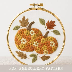 May include: A circular embroidery hoop containing white fabric with a hand-stitched design of two pumpkins composed of orange and yellow flowers, surrounded by embroidered brown and green leaves. The text PDF EMBROIDERY PATTERN is visible.
